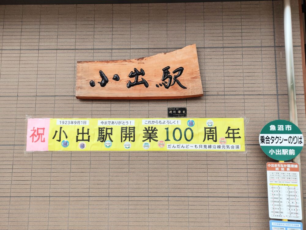 小出駅の壁に掲げてある、崩した字体で「小出駅」と書かれた木造の看板と、その下の「祝小出駅開業100周年」と書かれた横断幕の画像
